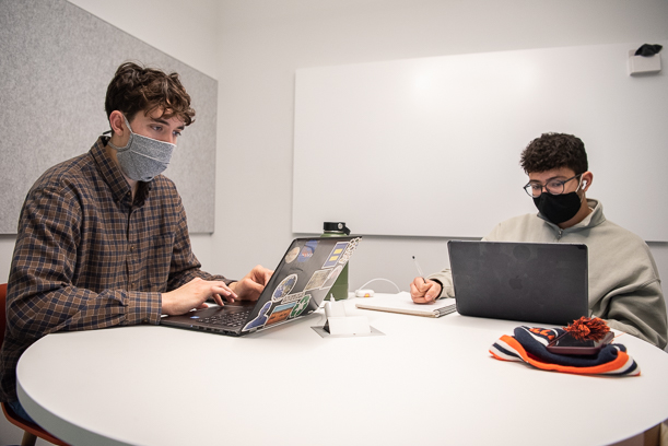 Two masked patrons sit at a white study room table where they are looking at computers.